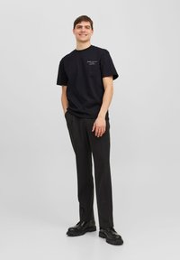 Camiseta de algodón negra de manga corta, con impresión de texto blanco. Combinada con pantalones negros y zapatos negros gruesos. El modelo está de pie con las manos en los bolsillos.