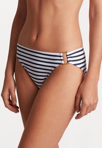 Μπικίνι κάτω μέρος με ναυτικό και λευκές ρίγες σε χαμηλή σχεδίαση, διακοσμημένο με χρυσές λεπτομέρειες στις πλευρές. Λείος υφασμάτινος υφή.