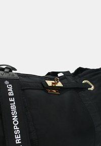 Zwarte nylon tas met een gouden klepsluiting, voorzien van een opvallende witte strap met het label "VERANTWOORDE TAS." Gladde textuur en gestructureerde vorm.
