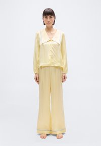 Γυναίκα όρθια, ξυπόλυτη, φορώντας απαλό κίτρινο σατέν σετ πιτζάμες με φαρδιά γιακά και μπλε λεπτομέρειες, σε λευκό απλό φόντο.