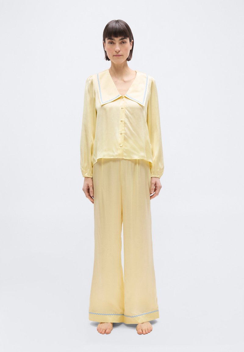 Γυναίκα όρθια, ξυπόλυτη, φορώντας απαλό κίτρινο σατέν σετ πιτζάμες με φαρδιά γιακά και μπλε λεπτομέρειες, σε λευκό απλό φόντο.