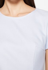 Světle modrý top s krátkým rukávem, vyrobený z texturované tkaniny, s kulatým výstřihem a detailem kapsy na přední straně. Hladký povrch a přiléhavý střih.