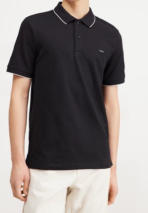 Czarna koszulka polo z krótkim rękawem, białą lamówką na kołnierzyku i rękawach, z zapięciem na dwa guziki, noszona z jasnymi spodniami.