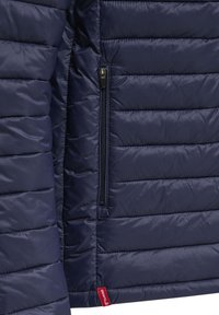 Marineblaue gesteppte Jacke mit horizontalen Nähten, einer vertikalen Reißverschlusstasche und einem kleinen roten Markenschild nahe dem Saum.