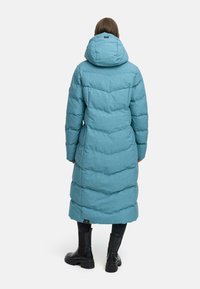 Lang puffjakke i lys blå, med hette, vertikal quiltet design og svarte detaljer, sammen med svarte støvler.