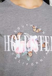 Grå bomullströja med blommönster, rosa blommor och orange fjärilar, accentuerad med texten "HOLLISTER SOUTHERN CALIFORNIA."