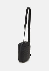 Bolso cruzado negro con tela texturizada que presenta un patrón de logo repetido. Tiene un diseño acolchado, una correa ajustable y un bolsillo lateral con cremallera.