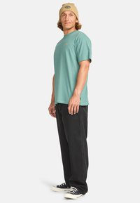 Camiseta de algodón verde menta con cuello redondo, combinada con vaqueros negros holgados y zapatillas azul marino. Los accesorios incluyen un gorro color canela con logo.