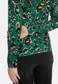 Langarmshirt in Grün mit einem schwarz-orangefarbenen Leopardenmuster, mit gerippter Textur und figurbetonter Passform. Kombiniert mit schwarzen Hosen.