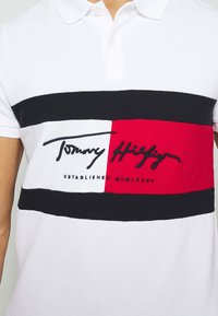 T-shirt polo blanc avec une bande horizontale noire, rouge et blanche ; présente le logo brodé "Tommy Hilfiger" et la date de création. Tissu doux.