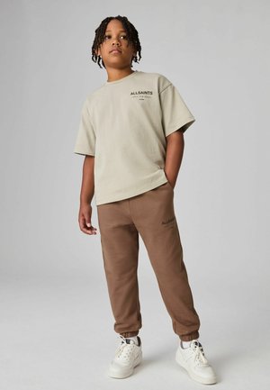 Lichtbeige T-shirt met korte mouwen met "ALLSAINTS" logo; bruine joggers met opgerolde enkels; witte sneakers; gladde katoenstructuur.