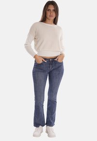 Maglione bianco a trama con scollatura ampia, abbinato a jeans slim-fit blu scuro e sneakers bianche. L'outfit presenta uno stile casual.