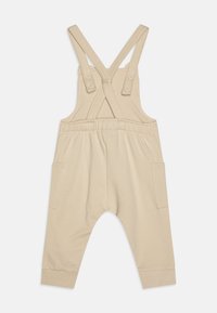 Beige bomuldsoveralls med krydsbagstropper, knaplukninger, elastisk talje og to sidelommer. Blød tekstur og tapered ben-design.