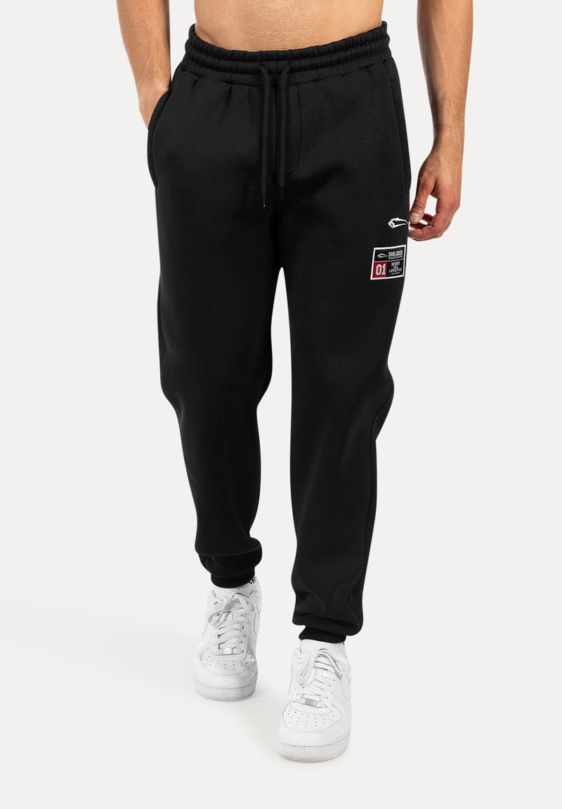 Schwarze Jogginghose mit Kordelzugbund, seitlichen Taschen und einem Logo-Patch mit roten und weißen Akzenten. Getragen mit weißen Turnschuhen.