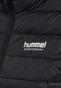 Zwart doorgestikte sportjas met zichtbaar wit hummel SPORTSWEAR-logo op de borst naast een zwarte rits.