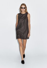 Bezramenné áčkové šaty z tmavě hnědé látky s hladkou strukturou, se bočními kapsami a kulatým výstřihem. Nosené s černými botami a slunečními brýlemi.