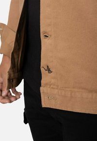 Chaqueta de algodón marrón con un ajuste holgado, que presenta un logo bordado negro en el dobladillo y detalles de costuras visibles. Botones cerca del cuello.