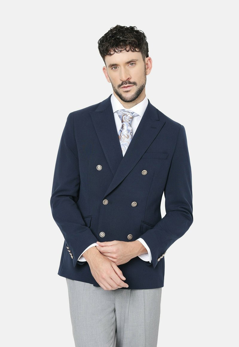Blazer azul marino de doble botonadura con botones metálicos, solapas de pico y bolsillos laterales; combinado con una camisa blanca y una corbata estampada.