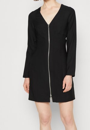 Woman wearing a black long-sleeve mini dress with a front silver zipper and V-neckline, standing against a plain light background.