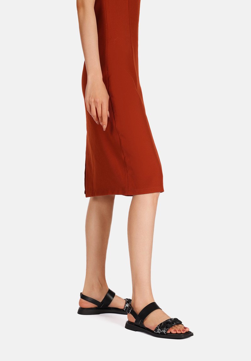 Sandali neri con suola piatta, caratterizzati da dettagli decorativi in tessuto nero e cinturini regolabili alla caviglia, abbinati a un vestito color ruggine.