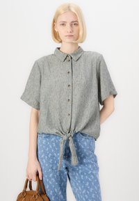 Camicia grigia con maniche corte e un nodo in vita, abbinata a jeans azzurri a fantasia e una borsa marrone.