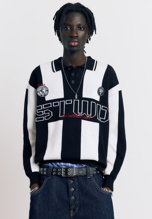 Jeune personne aux dreadlocks courtes portant un pull rayé noir et blanc avec un texte, et un jean en denim foncé, debout devant un fond uni.