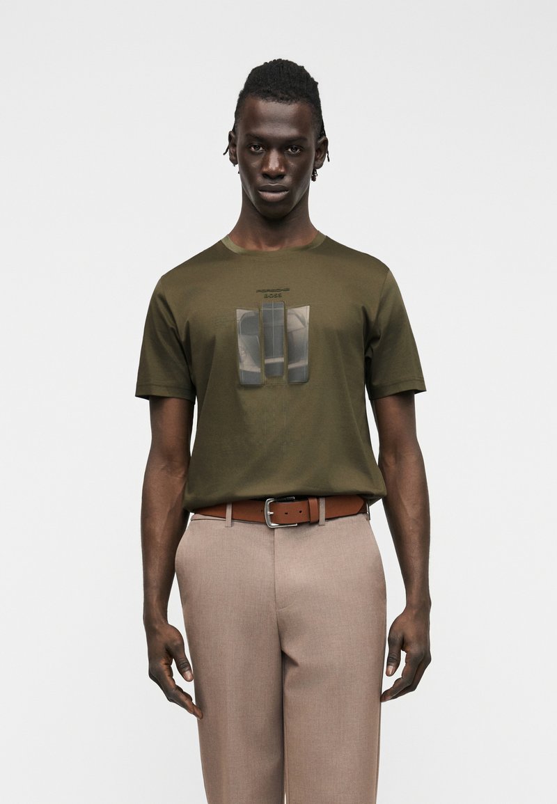 Άνδρας που φοράει ελιά πράσινη μπλούζα με τρεις αντανακλαστικές λωρίδες και μπεζ παντελόνι με καφέ ζώνη, στέκεται μπροστά σε λευκό φόντο.