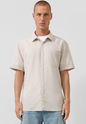 Mężczyzna o krótkich włosach, ubrany w jasnobeżową koszulę na krótki rękaw z guzikami na białą podkoszulkę i jasnoniebieskie dżinsy, stojący na tle szarego tła.