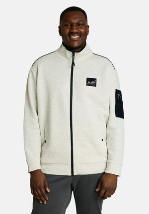 Smiling man wearing a light textured zip-up jacket with a small patch on the chest and a black zippered pocket on the left sleeve.