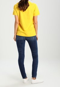 Žluté tričko s krátkým rukávem nošené s tmavě modrými skinny džíny a bílými teniskami. Materiál je hladký; střih je ležérní a pohodlný.