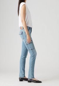 Mujer vistiendo jeans ajustados de color azul claro con bolsillos tipo cargo, una blusa sin mangas blanca y zapatos negros de tacón bajo, de pie de lado sobre un fondo liso.