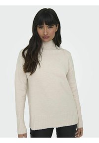 Pull col montant beige clair, texture tricotée, manches longues avec poignets côtelés, coupe décontractée, légèrement plus long à l'arrière, design minimaliste.