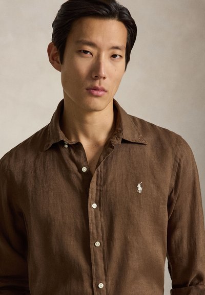 Tânăr cu păr închis la culoare poartă o cămașă maro cu nasturi, cu un mic logo alb brodat cu un cal pe piept.