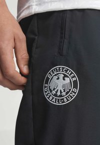 Κοντινή εικόνα ενός χεριού δίπλα σε μαύρο παντελόνι με λευκό λογότυπο αετού Deutscher Fussball-Bund κοντά στην τσέπη.