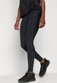 Person wearing black ruched top, black floral-patterned leggings, and black athletic sneakers against a plain light background.