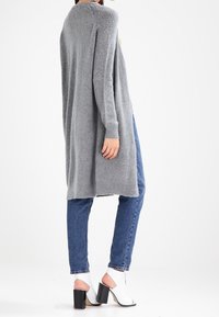 Ljusgrå lång cardigan i mjukt stickat material, med långa ärmar, öppen fram och avslappnad passform, tillsammans med blå jeans och vita klackar.