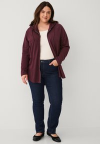 Frau mit braunen Haaren, die einen kastanienbraunen Reißverschluss-Hoodie, ein weißes Oberteil, dunkelblaue Jeans und schwarze Ballerinas trägt, steht da und hat eine Hand in der Tasche.