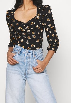 Femme portant un haut froncé noir à motifs floraux avec un décolleté carré et des manches bouffantes, associé à un jean taille haute bleu clair, les mains dans les poches.