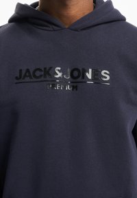 Person iført en mørkeblå hættetrøje med "JACK & JONES PREMIUM" i skinnende sort og sølv tekst på brystet.