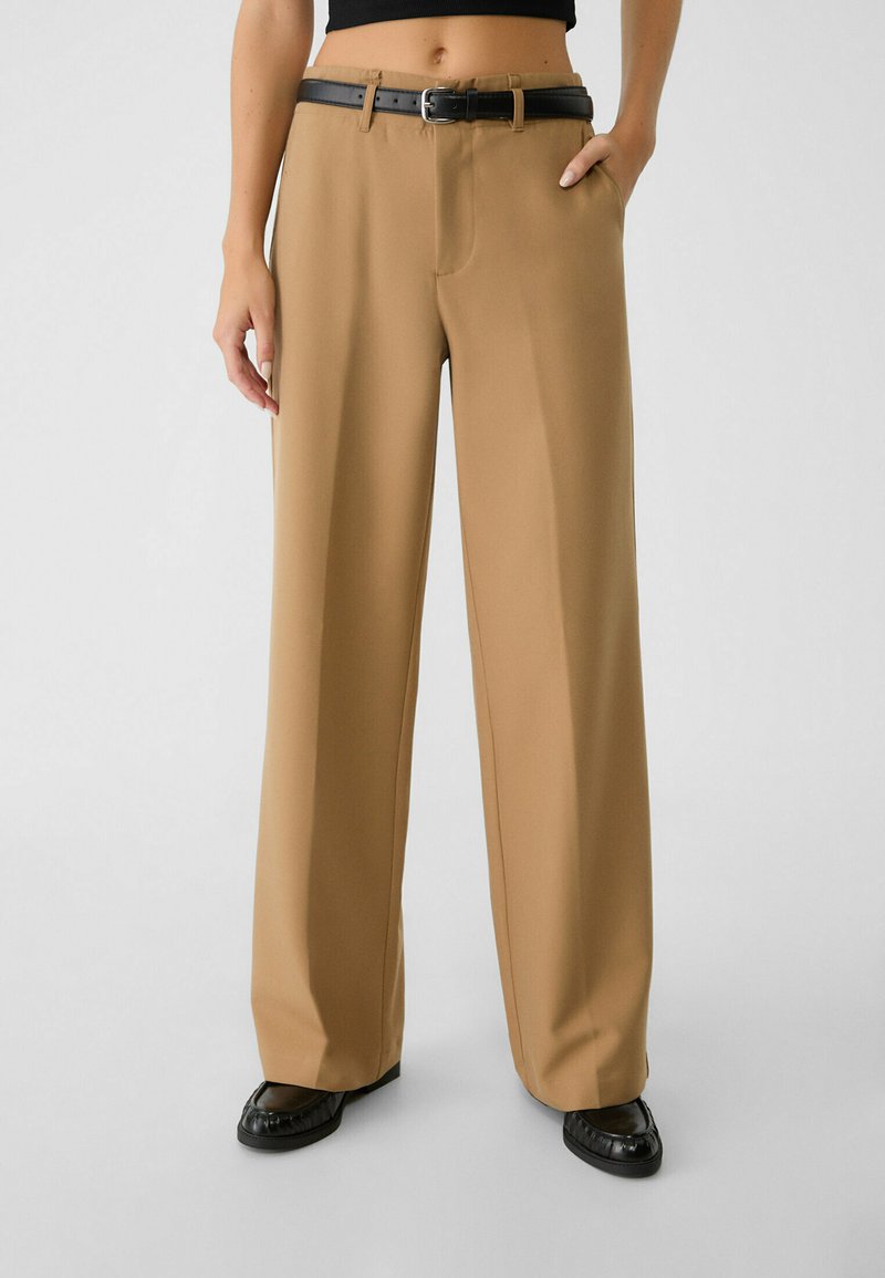 Pantaloni a gamba larga color beige realizzati in tessuto morbido, con una tasca frontale, una cintura nera in vita e calzature nere abbinate.