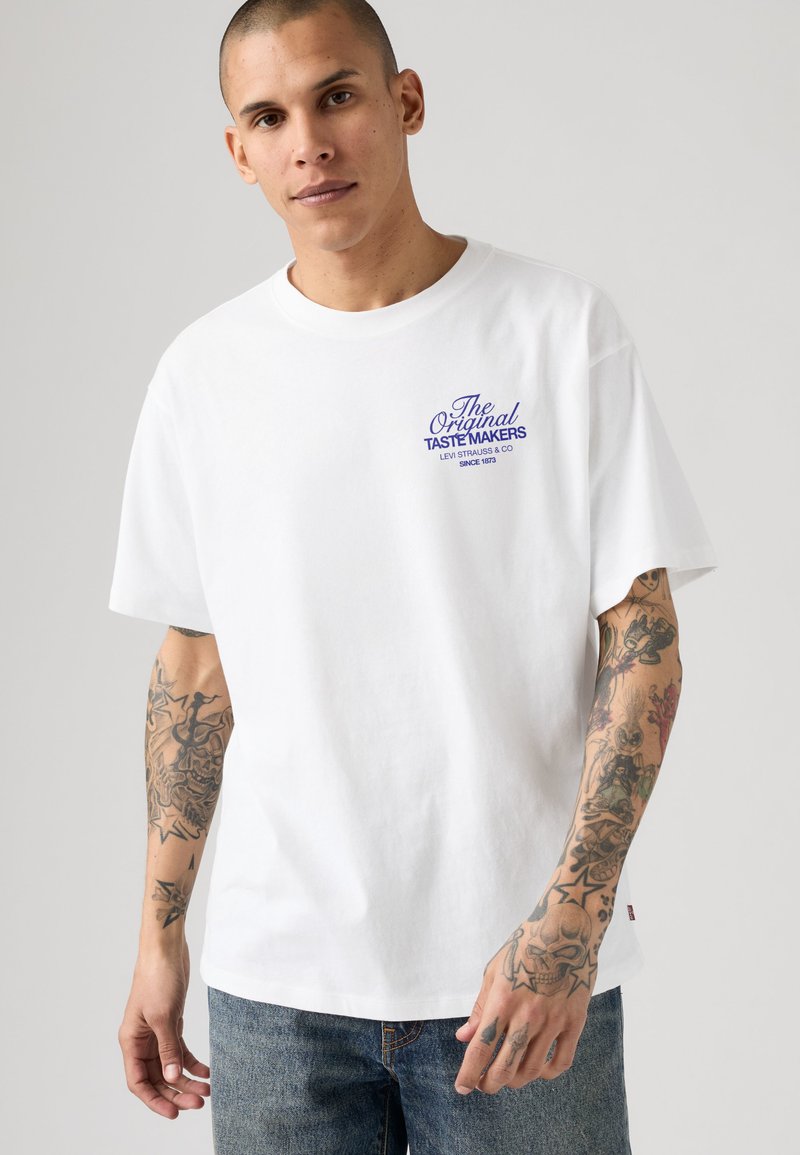 Giovane uomo con taglio rasato, indossa una maglietta bianca con scritta blu "The Original Taste Makers" e braccia pesantemente tatuate, in piedi contro uno sfondo uniforme.