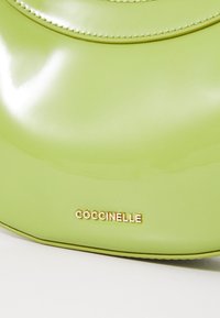 Vihreä lakattu nahkalaukku, jossa on kiiltävä pinta, kaareva muoto ja kultainen "Coccinelle" logo. Ompeleet tuovat lisää tekstuuria.