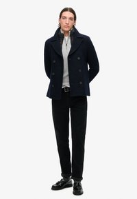Abrigo de lana azul marino tipo pea coat con frente de doble botonadura, gran cuello y acentos de botones. Suéter gris con cremallera debajo. Pantalones negros ajustados y zapatos.