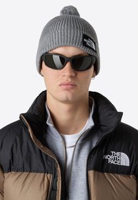 Šedá pletená čepice s bambulí, černé pilotní sluneční brýle a nadýchaná bunda s hnědými a černými sekcemi s nášivkou loga.