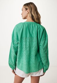 Maglietta verde a maniche lunghe con motivi ricamati a taglio, vestibilità rilassata e scollatura arricciata, abbinata a pantaloncini bianchi.