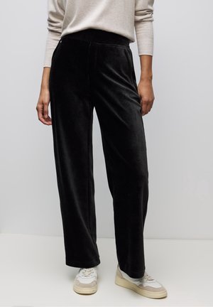 Person wearing black wide-leg velvet pants, beige long-sleeve top, and white sneakers against a plain background.