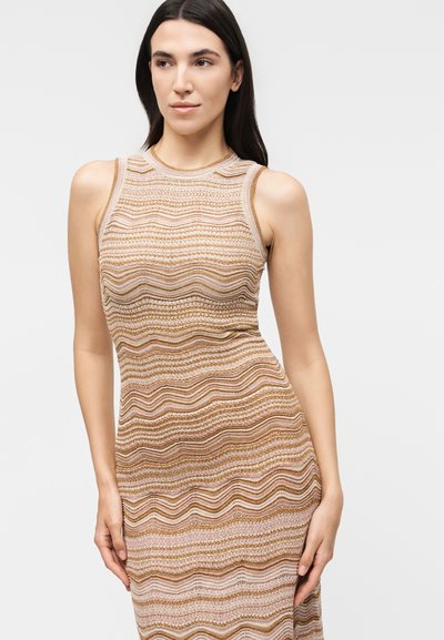 Γυναίκα με μακριά σκούρα μαλλιά, που φοράει ένα αμάνικο, εφαρμοστό φόρεμα με οριζόντιες κυματιστές ρίγες σε μπεζ, καφέ και ανοιχτό ροζ αποχρώσεις.