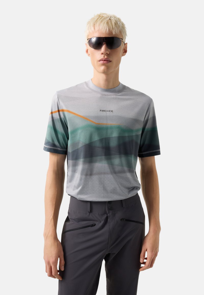 Junger Mann mit kurzen blonden Haaren, der dunkle Sonnenbrillen trägt, ein graues Grafik-T-Shirt mit dem Schriftzug "FIRE+ICE" und dunkle Hosen mit Reißverschlusstaschen.