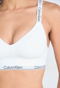 Witte bralette met een V-halsontwerp, voorzien van verstelbare bandjes en een band gemarkeerd met "Calvin Klein" in het zwart. Gladde textuur.