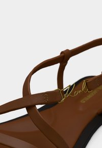 Καφέ δερμάτινο σανδάλι με λεπτές τιράντες και χρυσή λεπτομέρεια με το σήμα στο πάνω λουράκι, σε απλό λευκό φόντο.